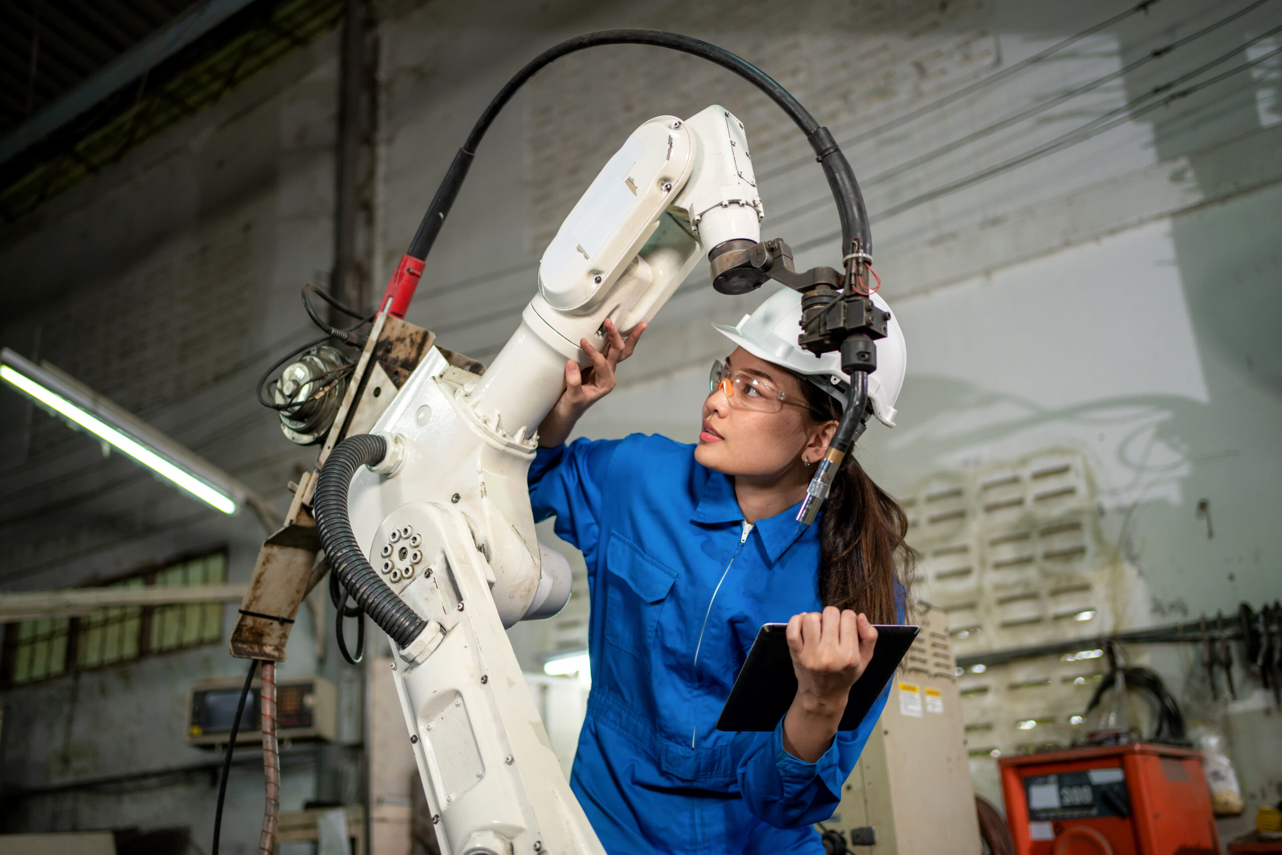Technician woman check the robotic arm machine in the factory. Worker wearing safety helmet, glasses and uniform. Preventive maintenance to prevent breakdown. Service and repair concept automation